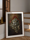 Framed artwork of a mistletoe hanging with a red ribbon on a wall.