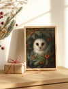 Framed artwork of an owl in a wreath on a wooden surface with a gift box.