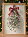 Framed artwork of mistletoe with a red bow on a neutral background, Christmas tree in the background.